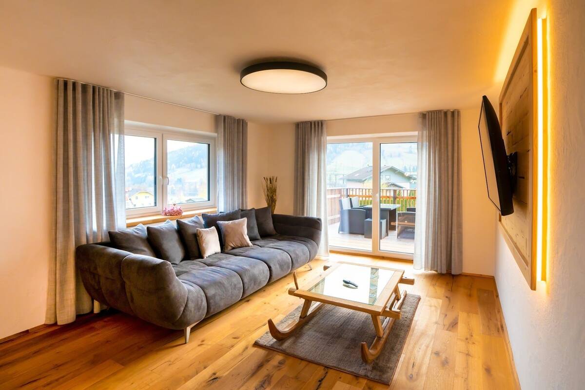 A modern living room with a grey sofa, a glass coffee table, wooden flooring, and large windows offering a view of a balcony with outdoor furniture.