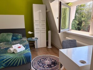 Een slaapkamer met een bed, een bureau, een stoel, een radiator, een raam met uitzicht op groen en een witte inloopkast.