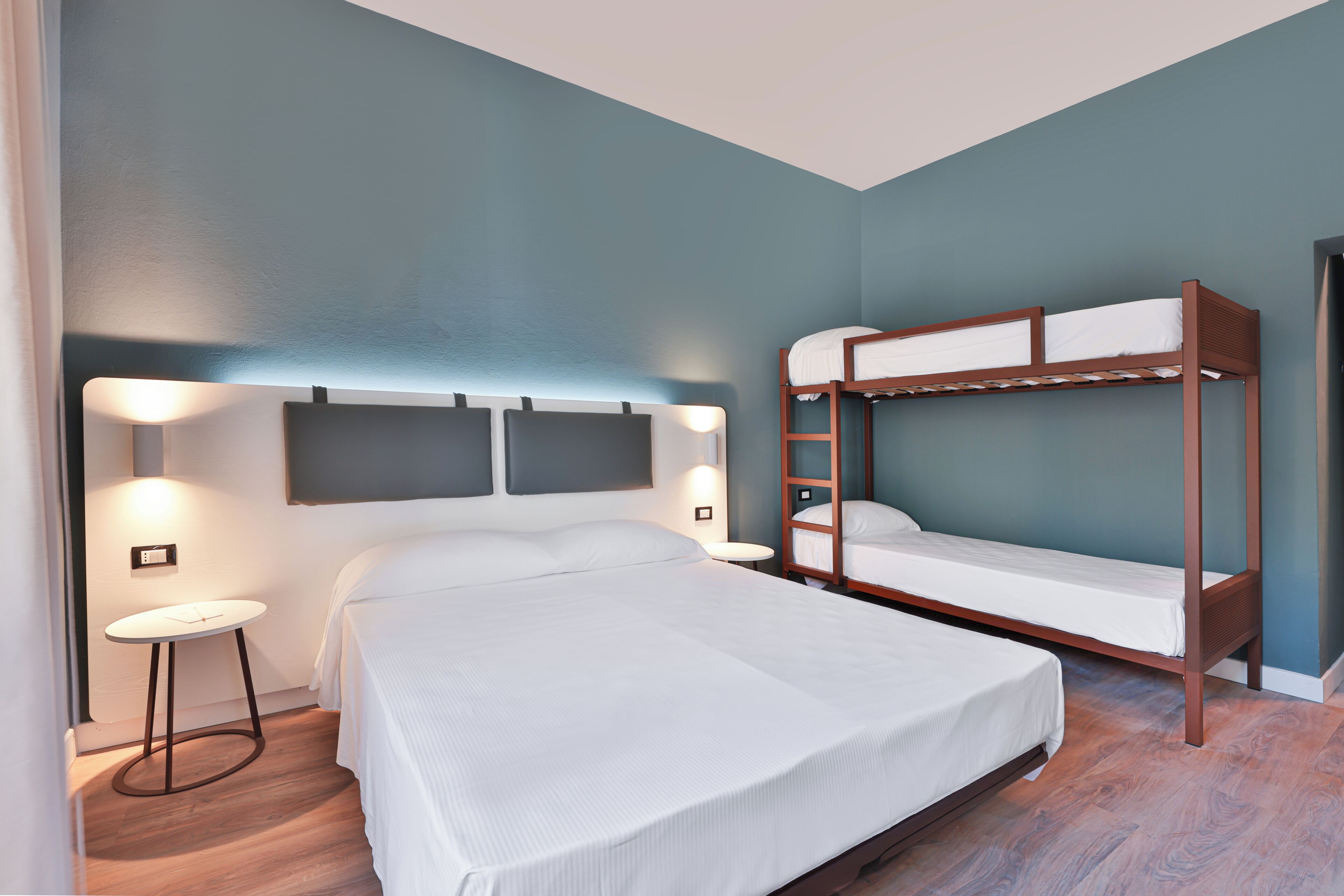 A bedroom with a large bed, a smaller bunk bed, a bedside table, and wall-mounted lighting.