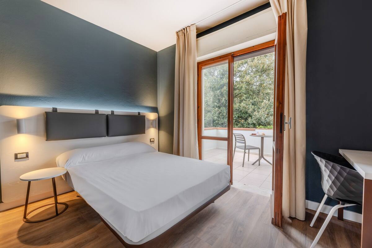 Una habitación de hotel moderna con una cama grande, un escritorio y un balcón con mobiliario exterior.