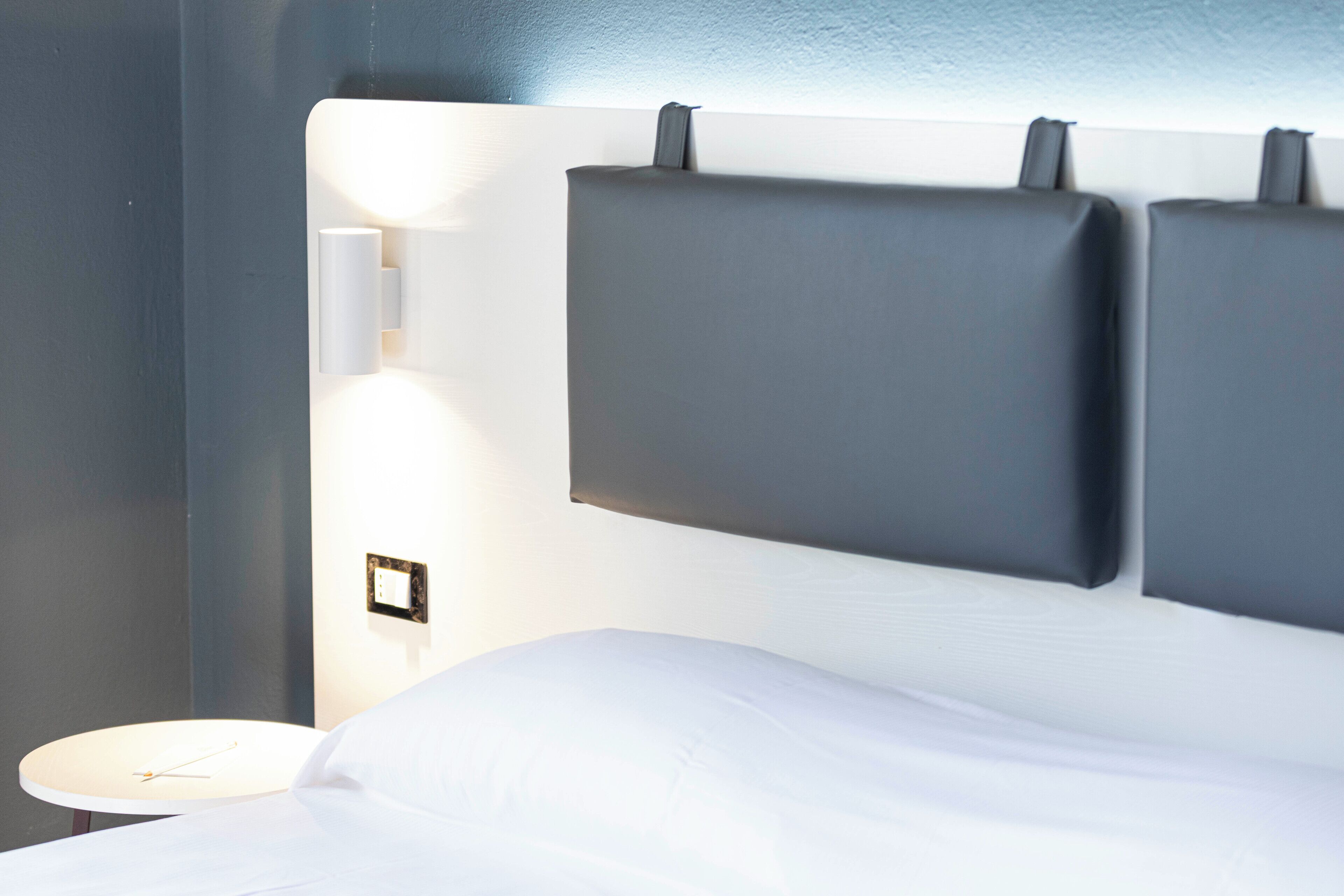 A modern hotel room with a large bed, a bedside table with a clock, and a wall-mounted light fixture.