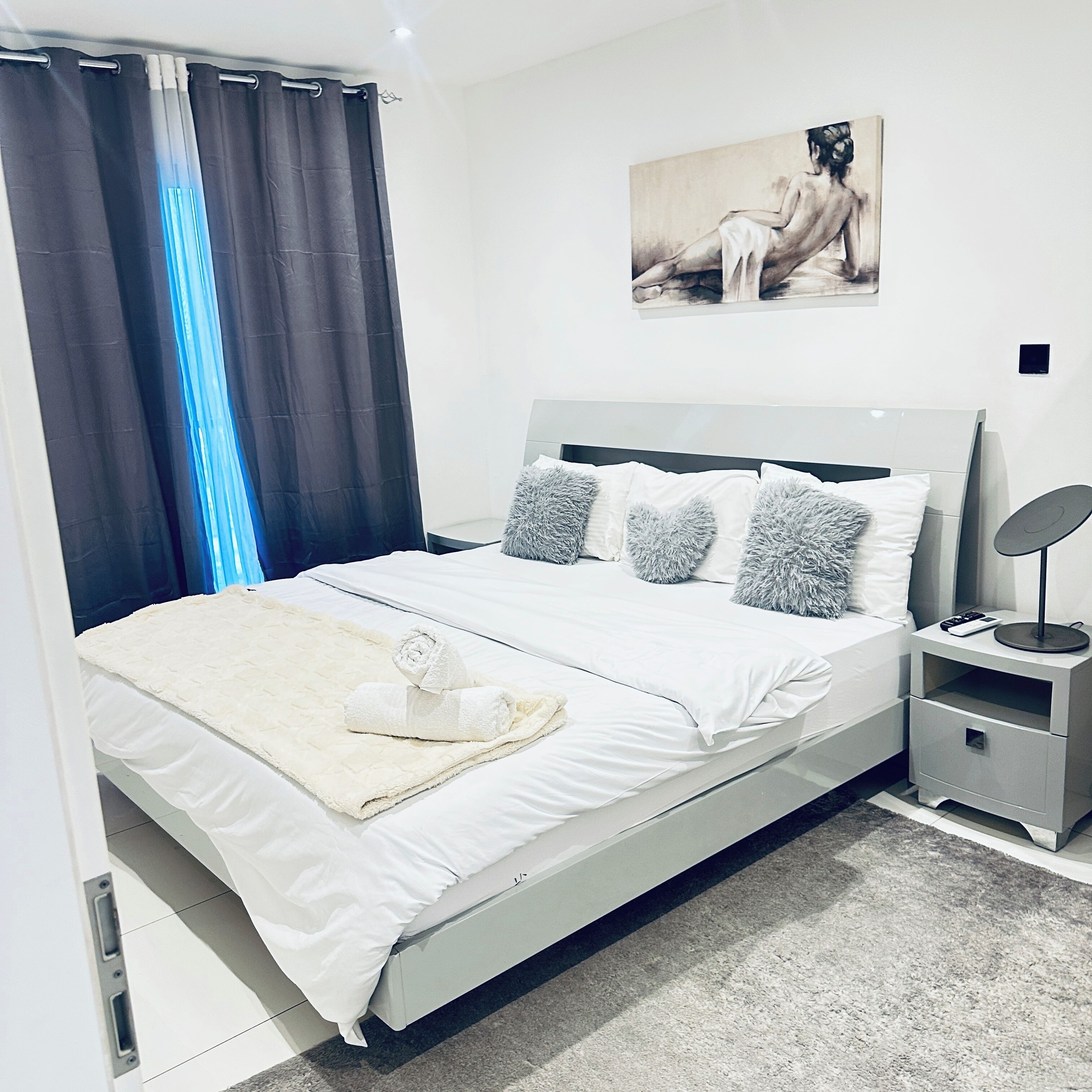 A modern bedroom with a bed, pillows, a blanket, a painting, and a nightstand.