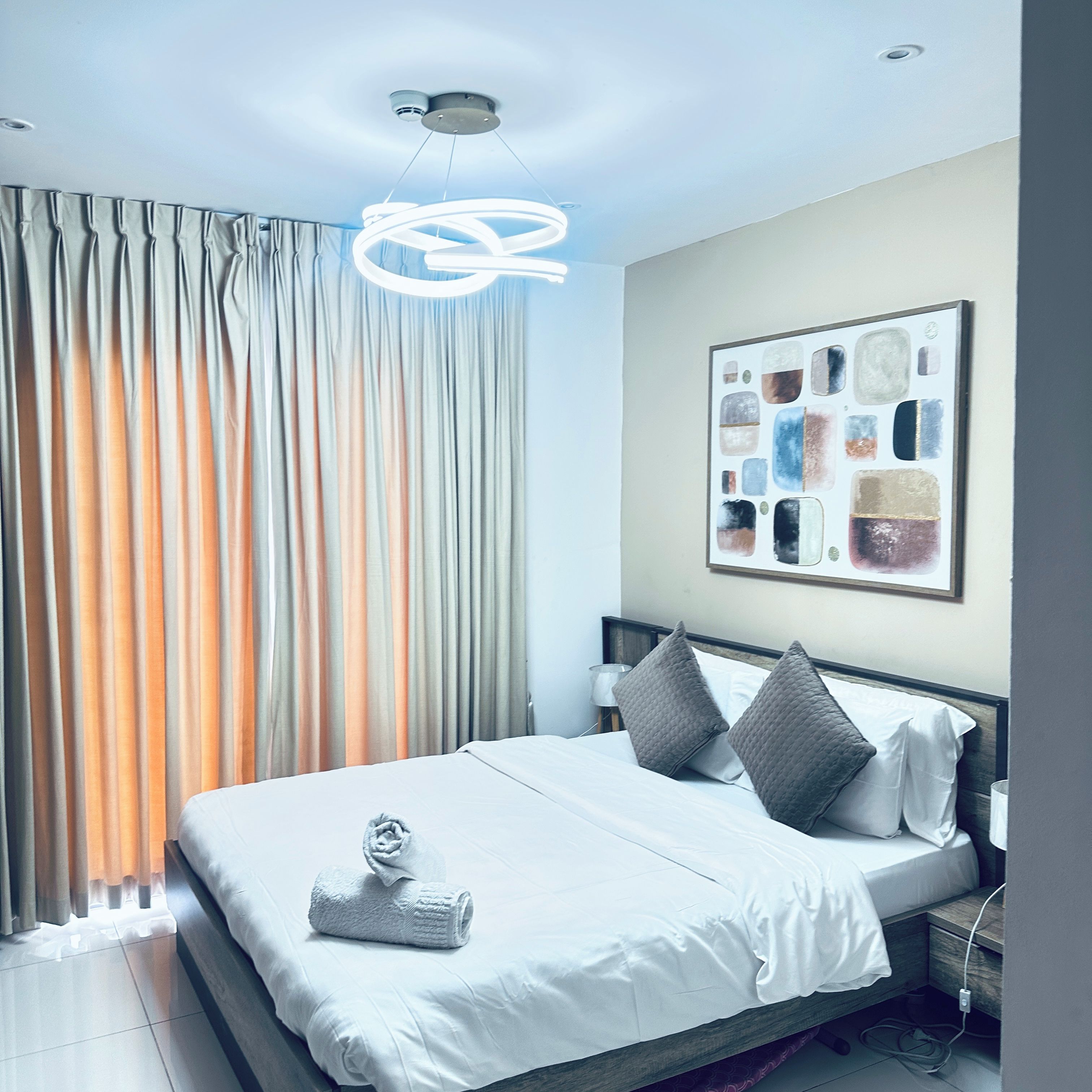 A modern hotel room with a neatly made bed, a wall-mounted abstract art piece, and a ceiling light fixture.