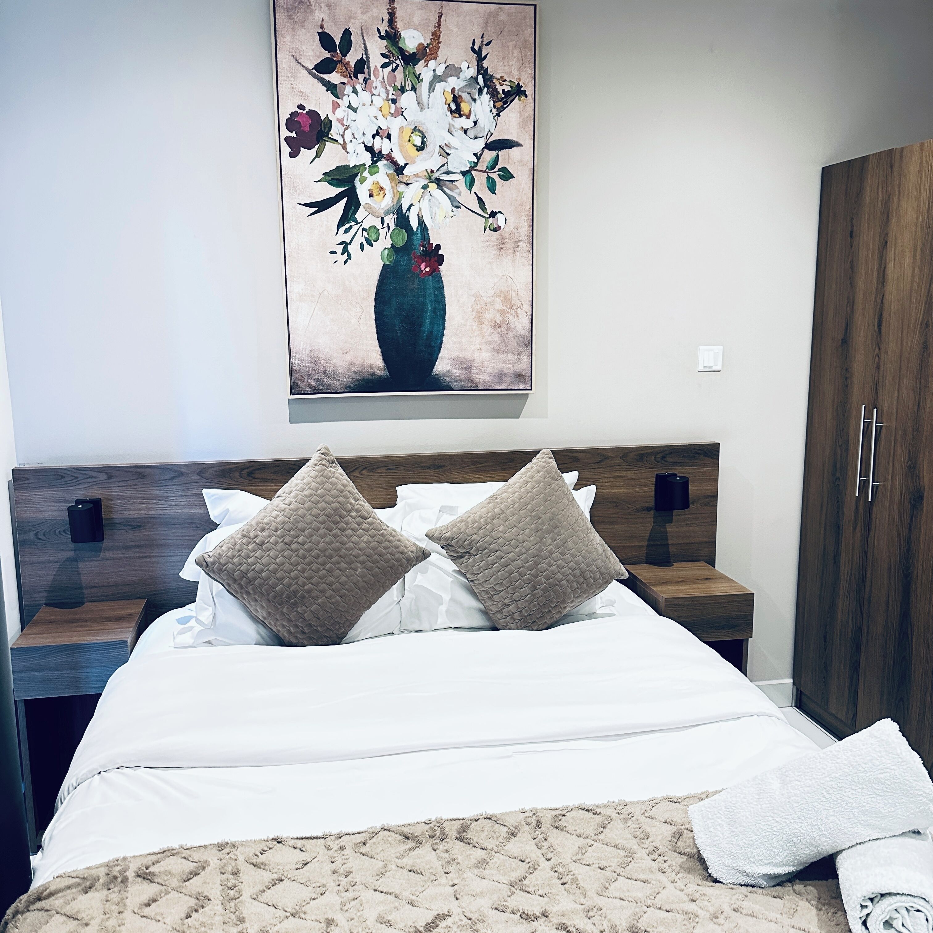 A neatly made bed with two pillows, a wall-mounted painting of flowers, and a wooden wardrobe.
