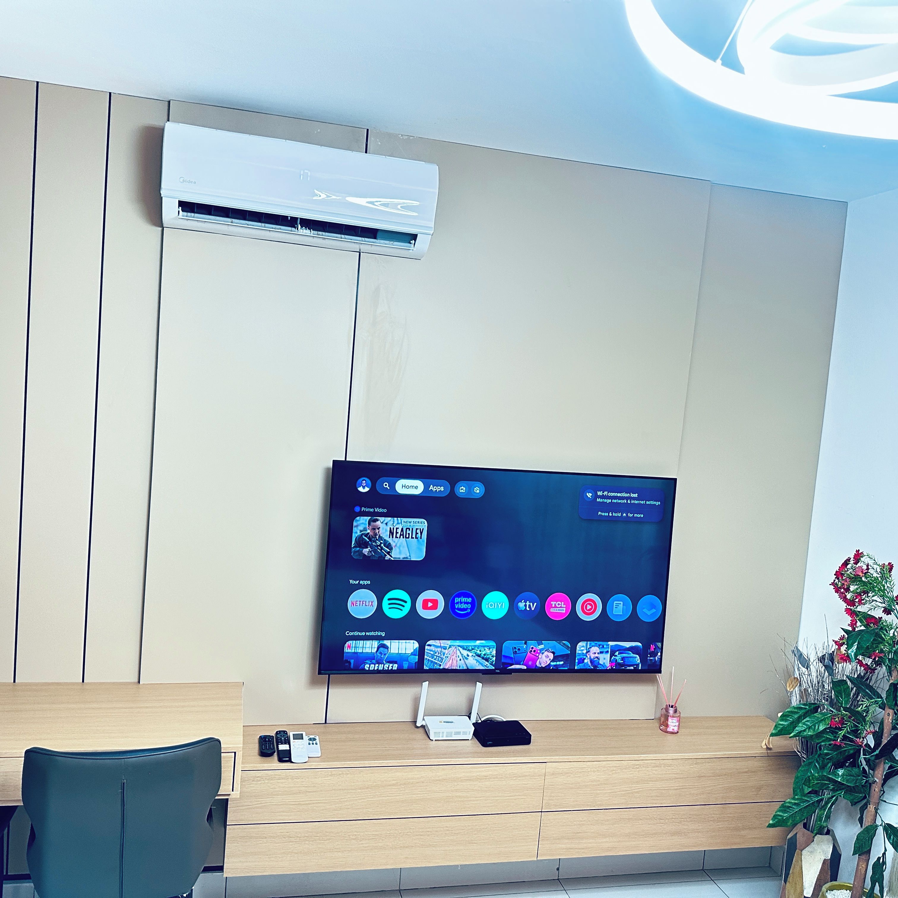 A modern living room with a flat-screen TV mounted on a wall, a built-in air conditioner, and a wooden coffee table with a laptop and a plant.