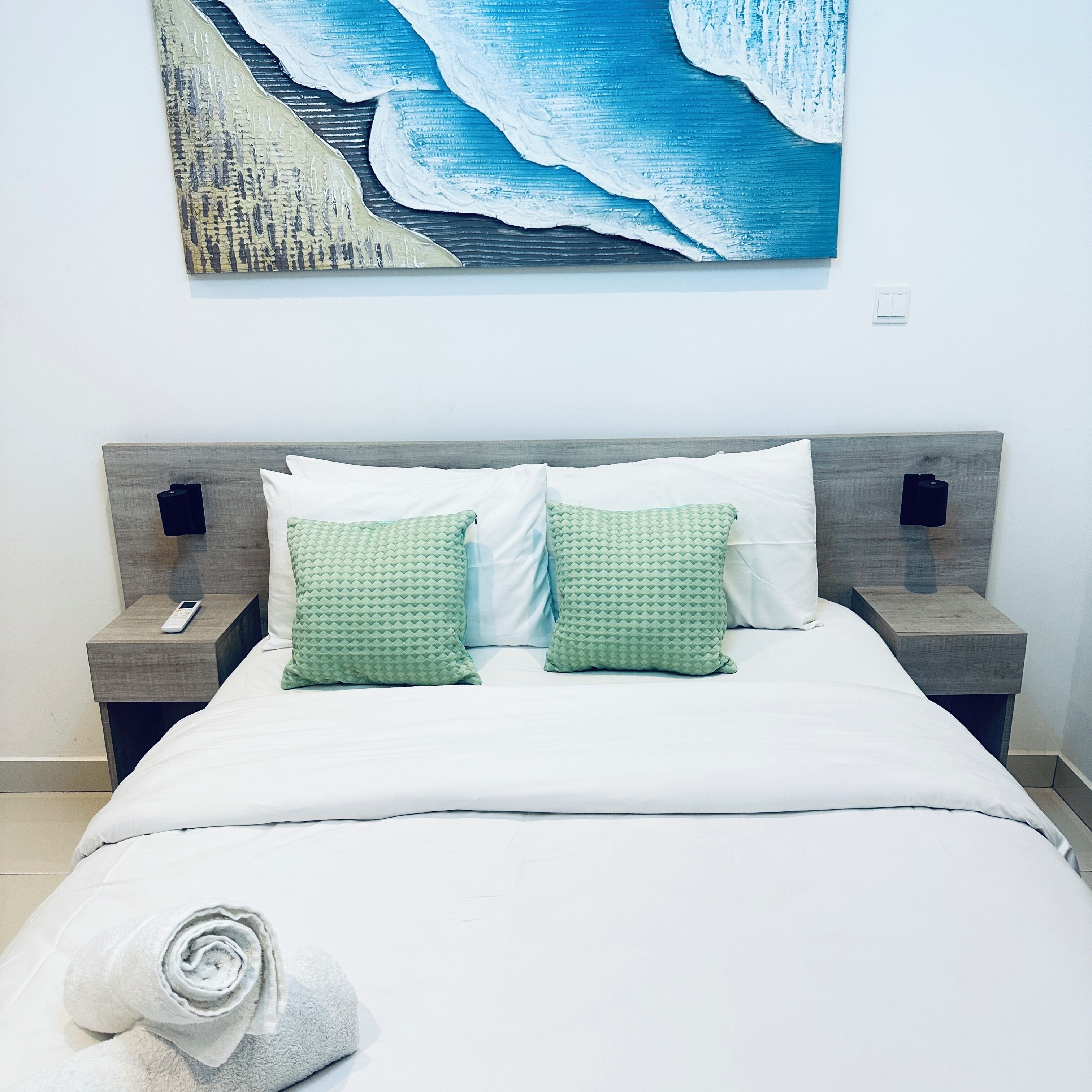 Un letto rifatto con lenzuola bianche e due cuscini verdi, un telecomando sul comodino e un quadro raffigurante un paesaggio marino sulla parete.