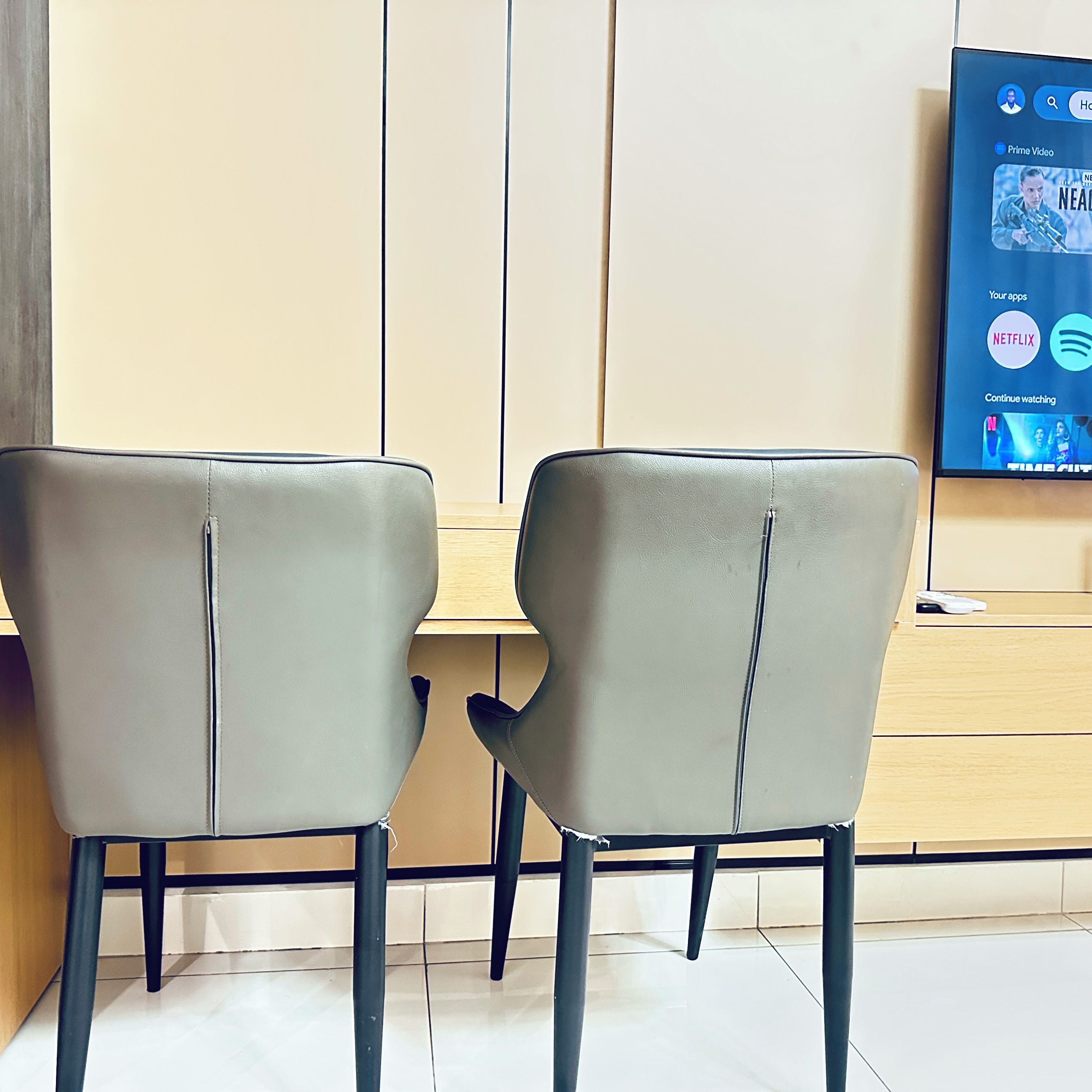 Dos sillas grises con patas negras, una mesa de madera y un televisor fijado a la pared mostrando la interfaz de una plataforma de streaming.