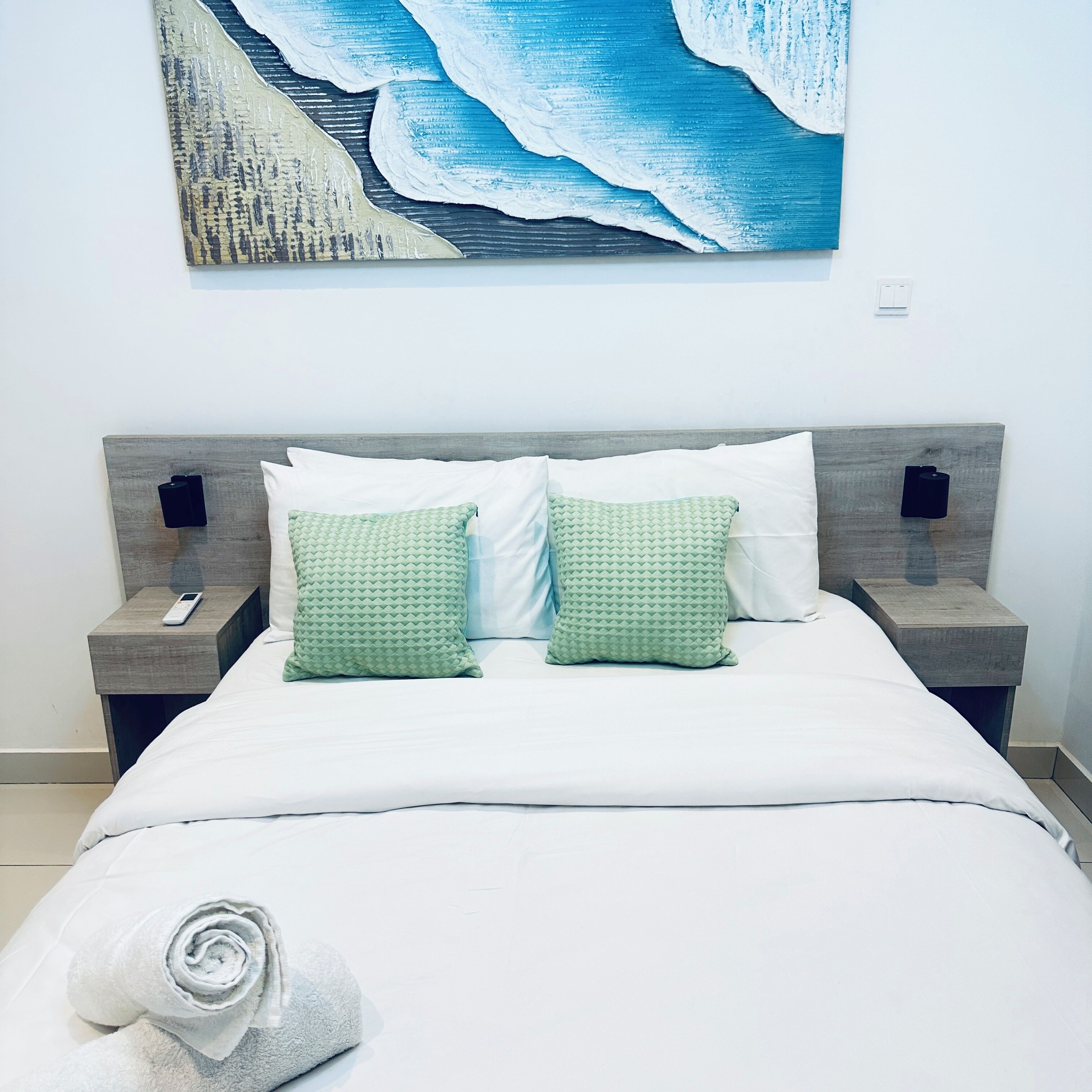 Un lit bien fait, avec des draps blancs, deux coussins à motifs verts, une télécommande sur une table de chevet et un tableau représentant un paysage marin bleu au-dessus du lit.