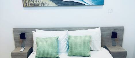 Un lit bien fait, avec des draps blancs, deux coussins à motifs verts, une télécommande sur une table de chevet et un tableau représentant un paysage marin bleu au-dessus du lit.