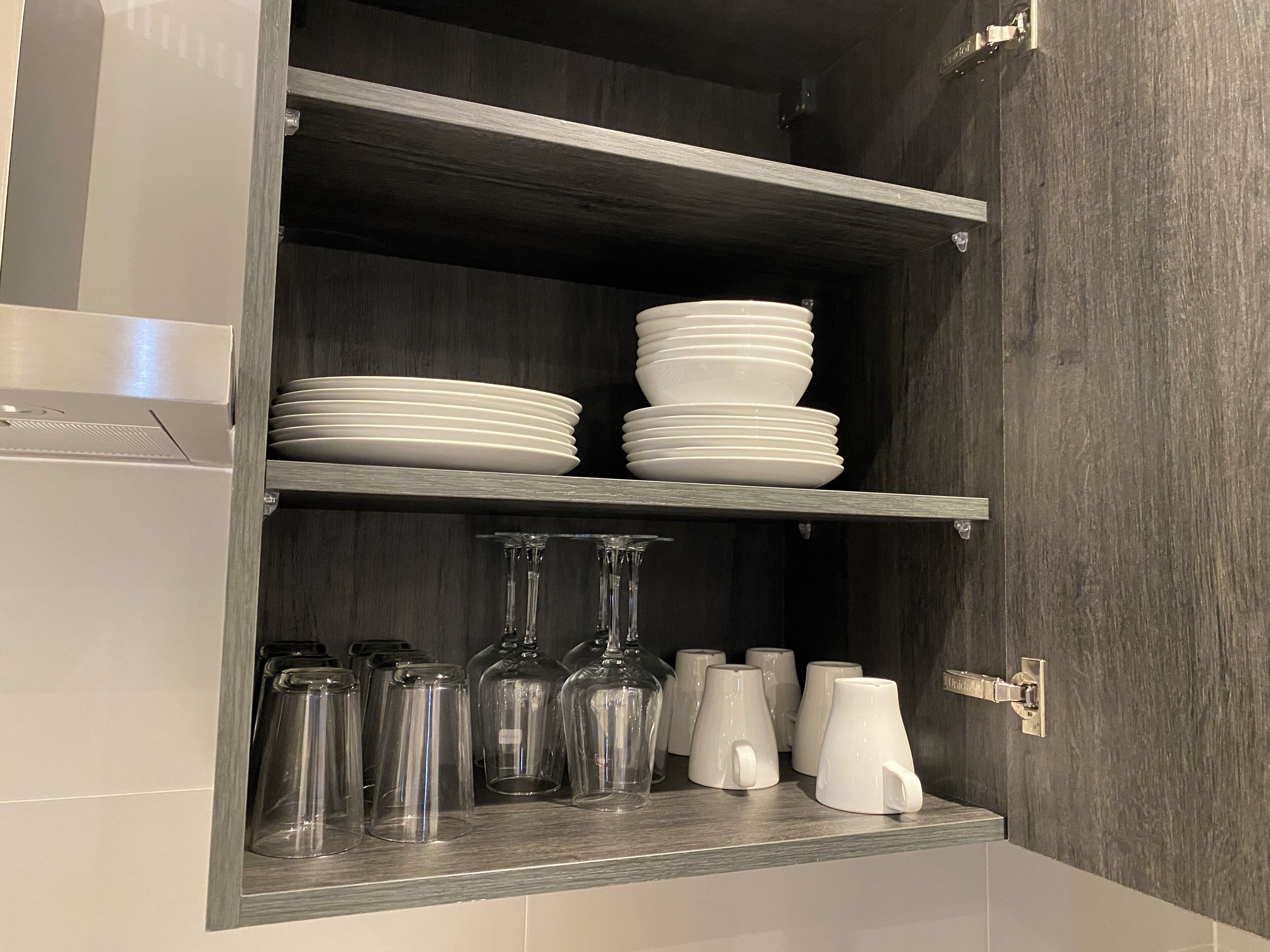 Un meuble de cuisine avec des étagères où sont rangées des assiettes blanches, de la verrerie transparente et des tasses blanches.