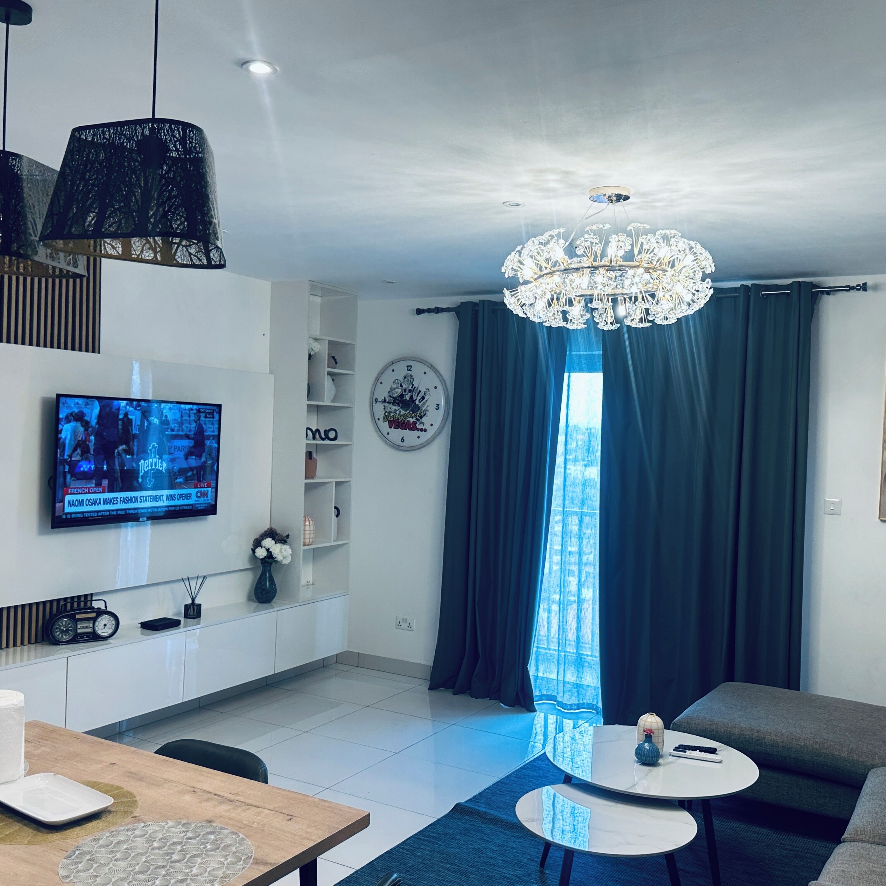 A modern living room with a flat-screen TV, a chandelier, a sofa, and a dining area with a table and chairs.