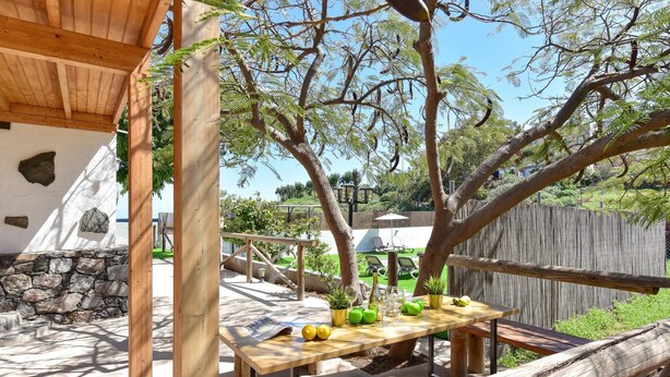An outdoor patio with a wooden table set for dining, surrounded by trees and a stone wall.