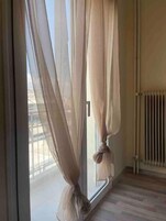 Quarto com cortinas translúcidas amarradas na parte inferior, um radiador na parede e piso de madeira.