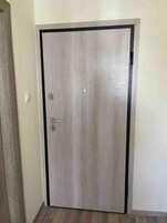 Uma porta fechada com puxador metálico e fechadura, embutida numa parede lisa com soalho de madeira.