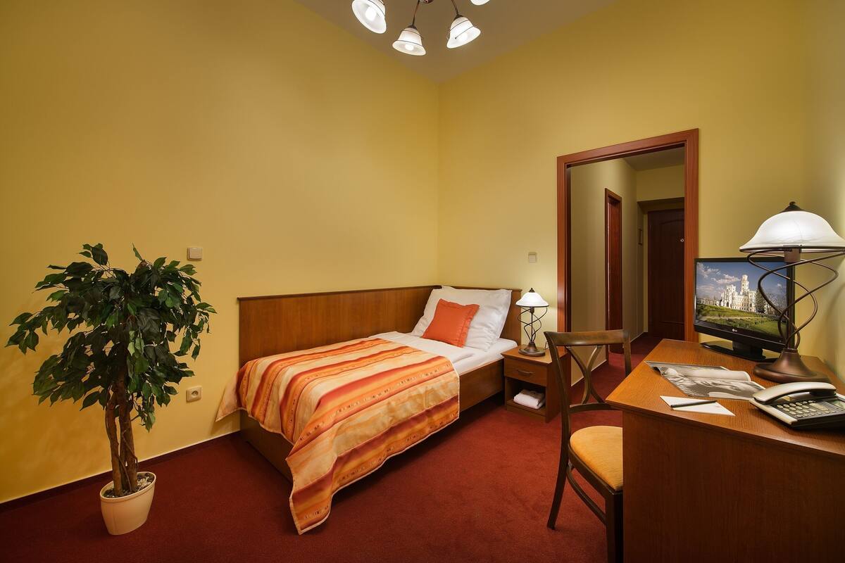 A hotel room with a bed, a desk with a computer, a lamp, a chair, and a potted plant.