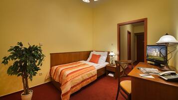 Quarto de hotel com uma cama, uma escrivaninha com um computador, um abajur, uma cadeira e um vaso de plantas.