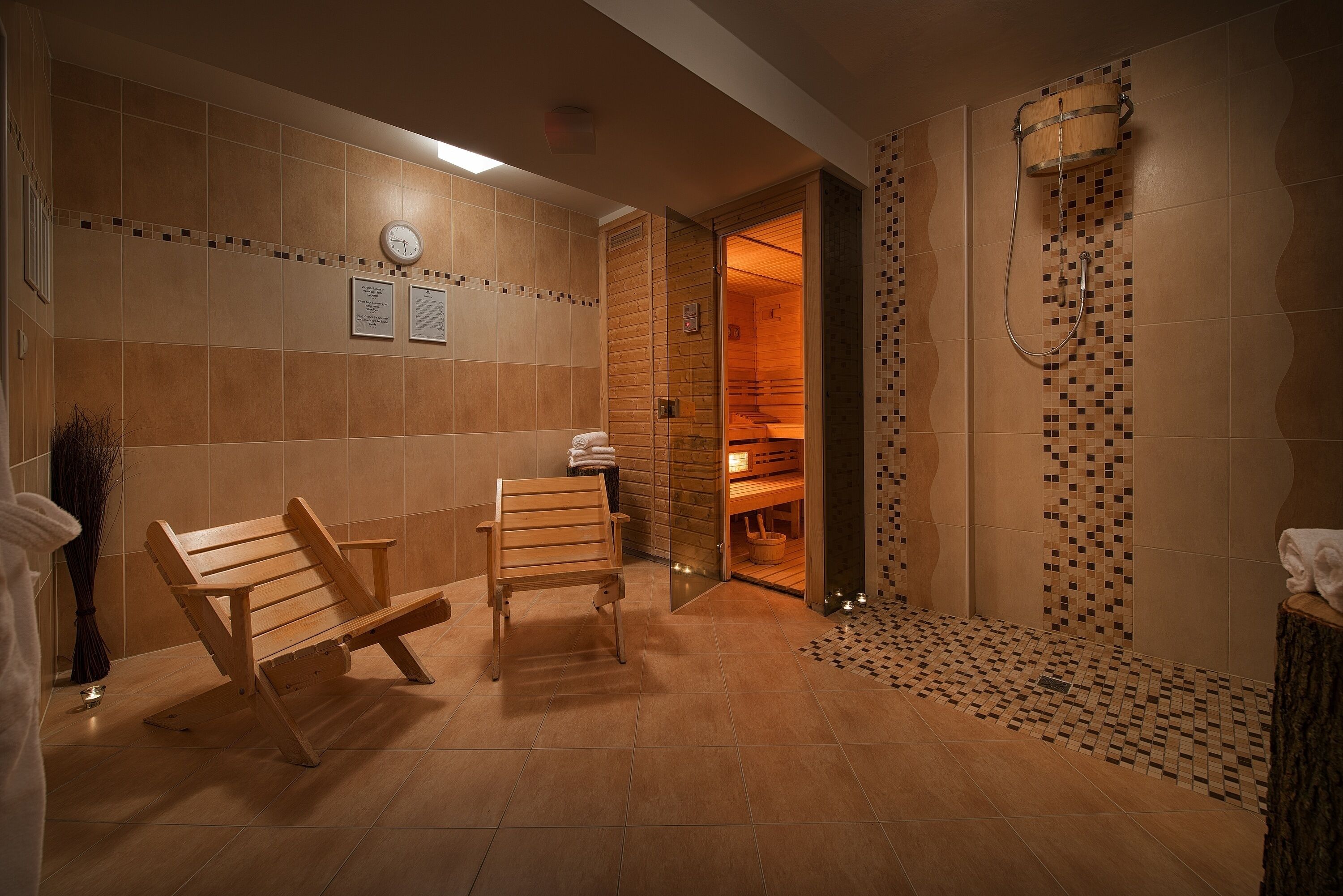 A sauna room with wooden benches, a shower area with a mosaic tile design, and a clock on the wall.