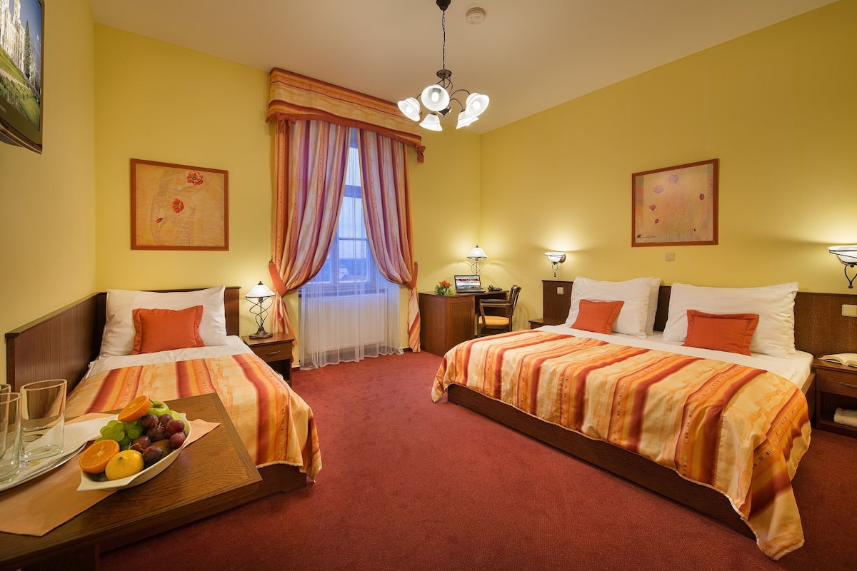 A hotel room with two beds, a large window with curtains, a chandelier, and artwork on the walls.