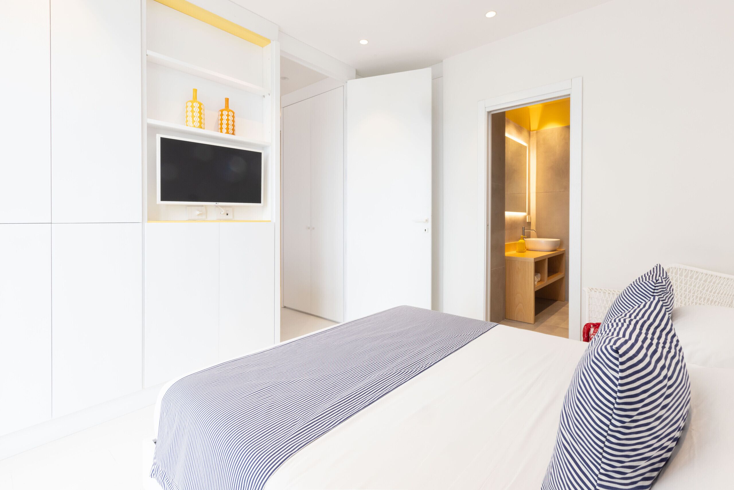 Quarto de hotel moderno com uma cama, televisão montada na parede e banheiro visível por uma porta aberta.