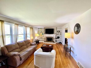 Un salon avec un canapé, un fauteuil blanc, une cheminée, un téléviseur et une horloge au mur.