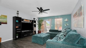 A modern living room with a flat-screen TV, a ceiling fan, a teal sofa, and a large window with curtains.