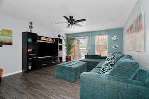 Un salon moderne doté d’un téléviseur à écran plat, d’un ventilateur de plafond, d’un canapé de couleur turquoise et d’une grande fenêtre avec des rideaux.
