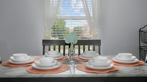 A dining table set with white plates and bowls, a vase with a single flower, and a black chair.