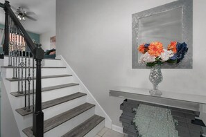 Un escalier avec une rampe noire et des marches en bois, menant à un palier agrémenté d’un miroir décoratif et d’un vase de fleurs.