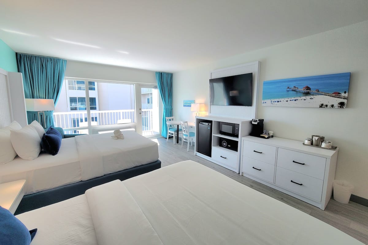 Une chambre d’hôtel avec un lit, un bureau avec une télévision, un balcon avec des rideaux et un tableau représentant une scène de plage.