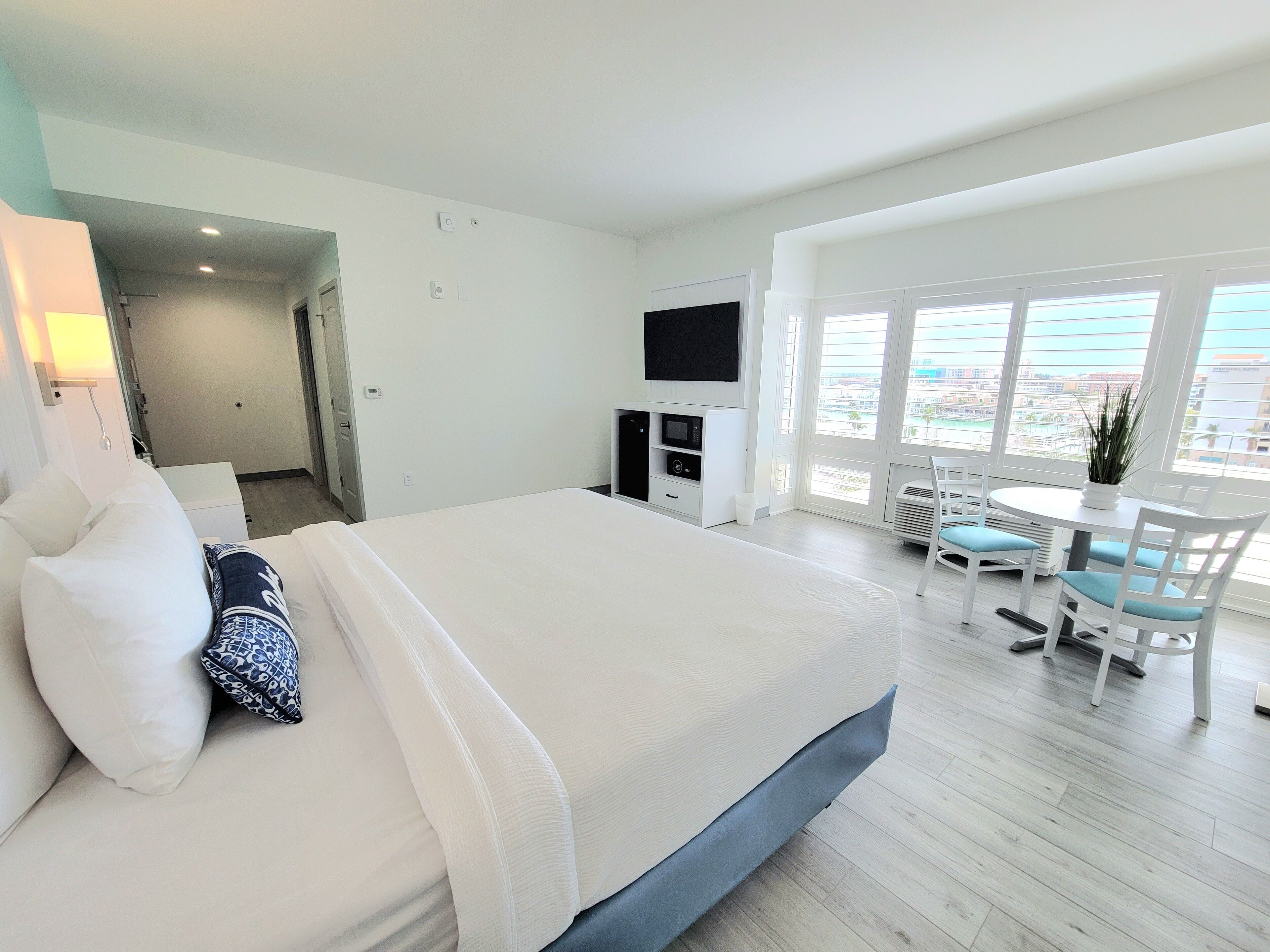 A hotel room with a bed, a TV, a dining area with chairs, and a view through large windows.