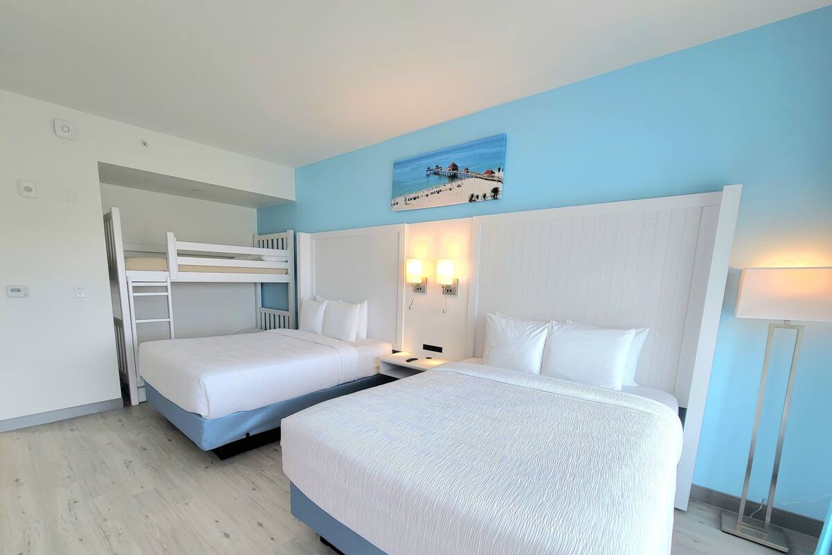 Une chambre d’hôtel avec deux lits, un lit superposé, un tableau représentant une scène de plage et une lampe de chevet.