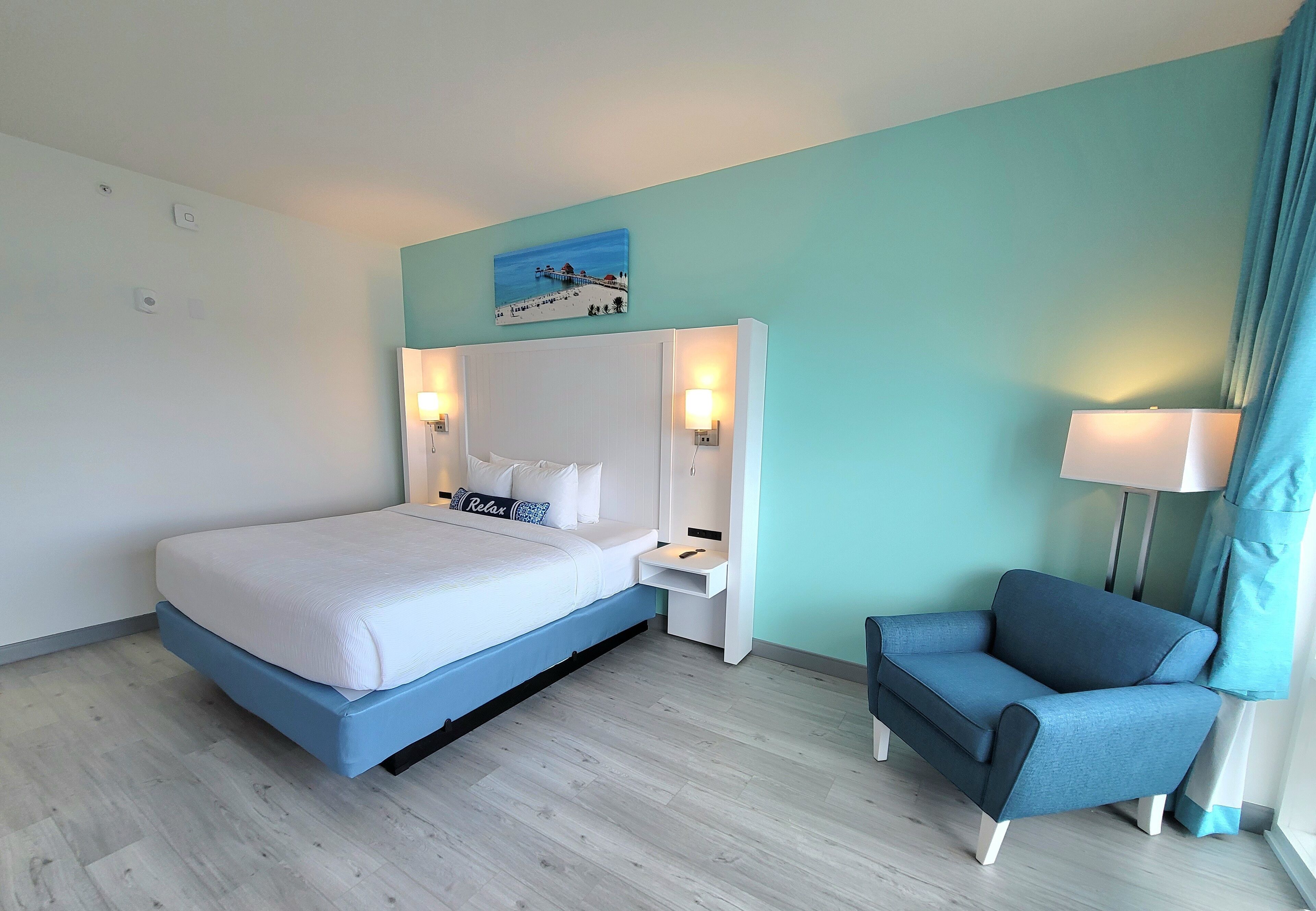 A modern hotel room with a bed, a nightstand, a chair, and a painting on the wall.