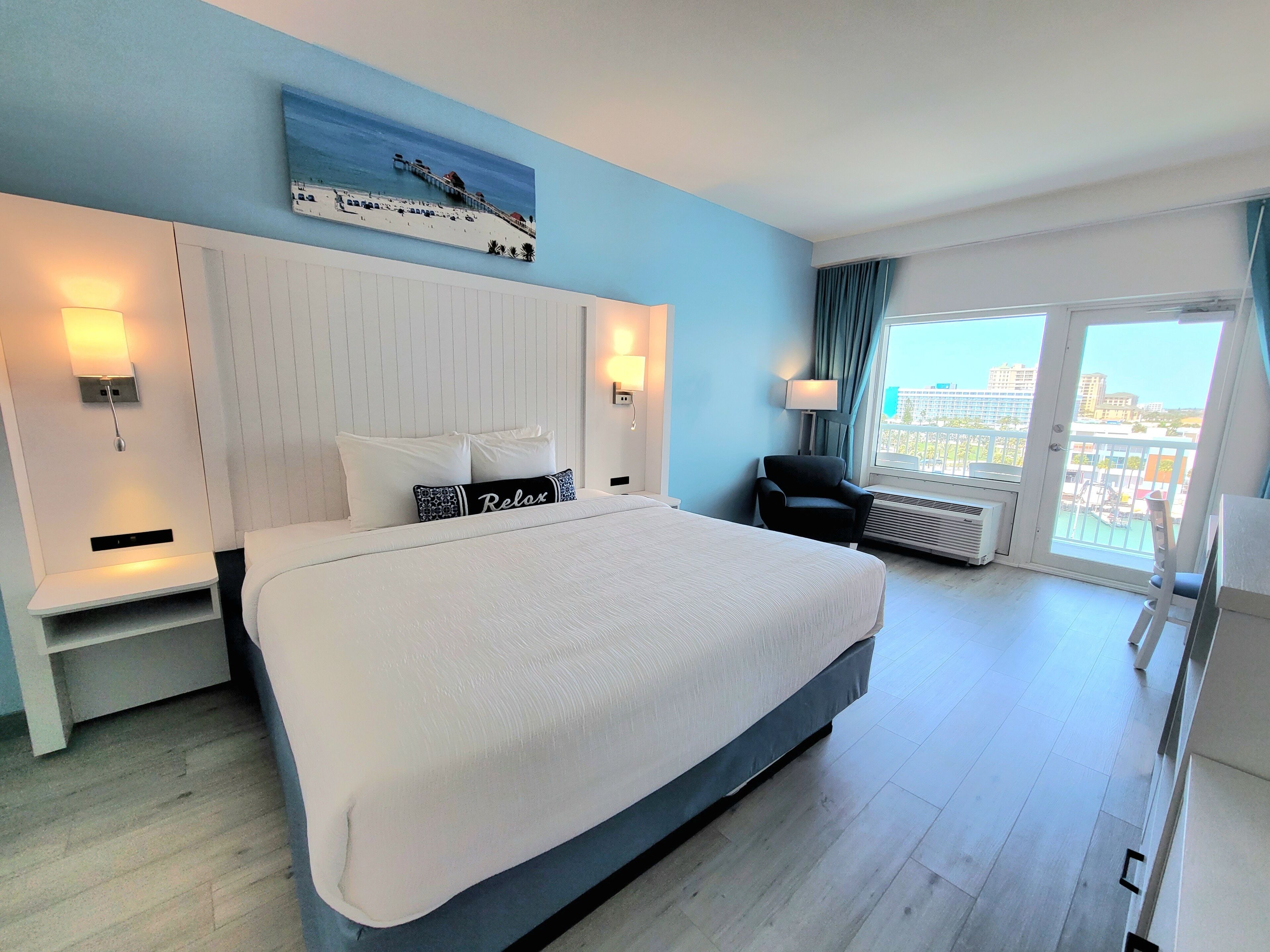 A hotel room with a large bed, a headboard with a cityscape painting, a chair, a window with a view of buildings, and a balcony with a railing.