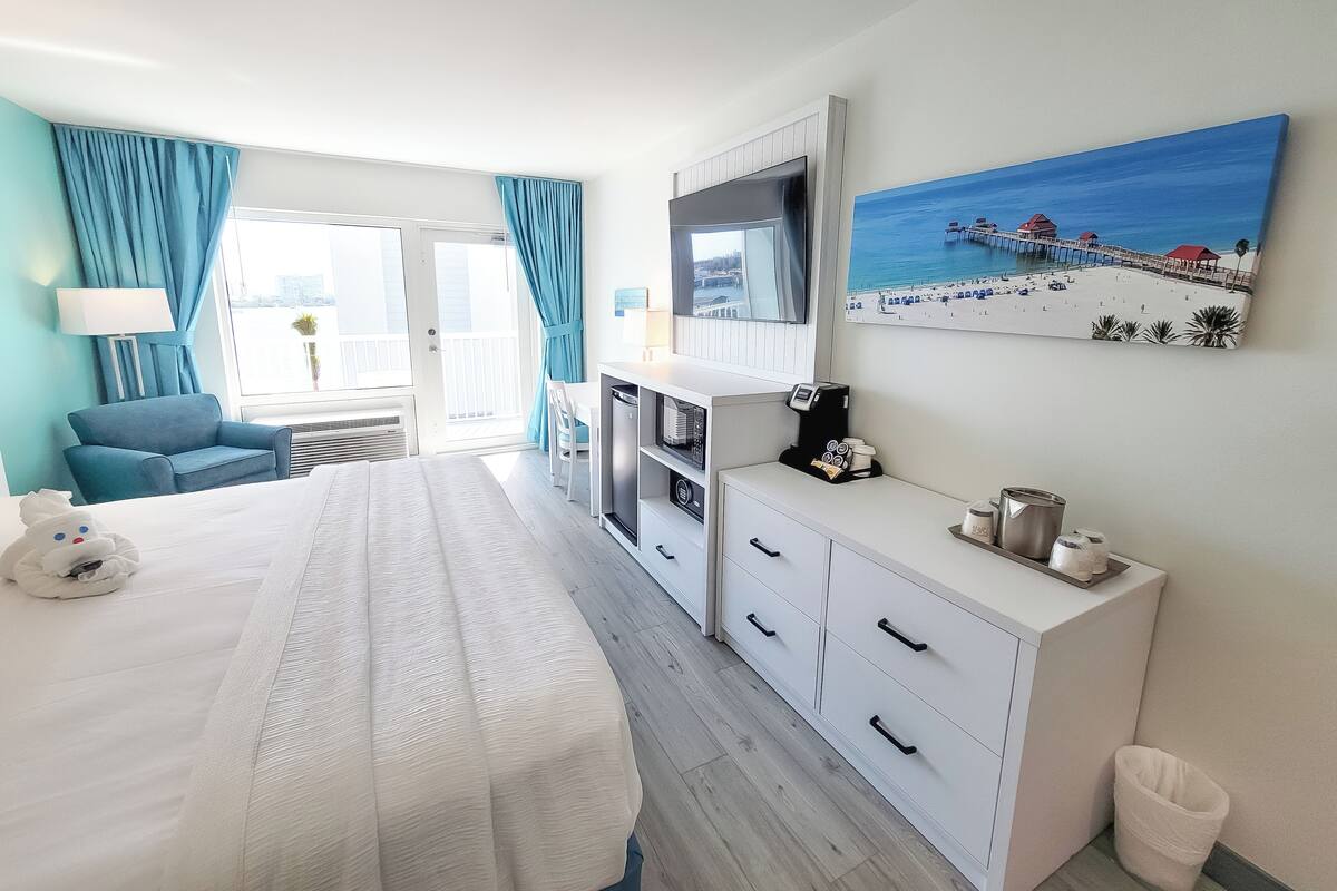 Une chambre d’hôtel équipée d’un lit, d’un bureau, d’une machine à café, d’une poubelle et d’un tableau représentant une scène de plage.