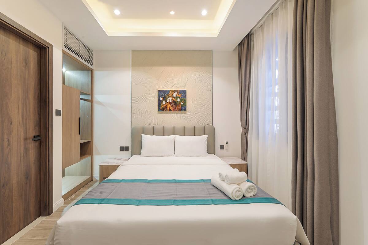 Ein Hotelzimmer mit Bett, Kopfteil, einem Bild an der Wand, einem Kleiderschrank und einem Fenster mit Vorhängen.