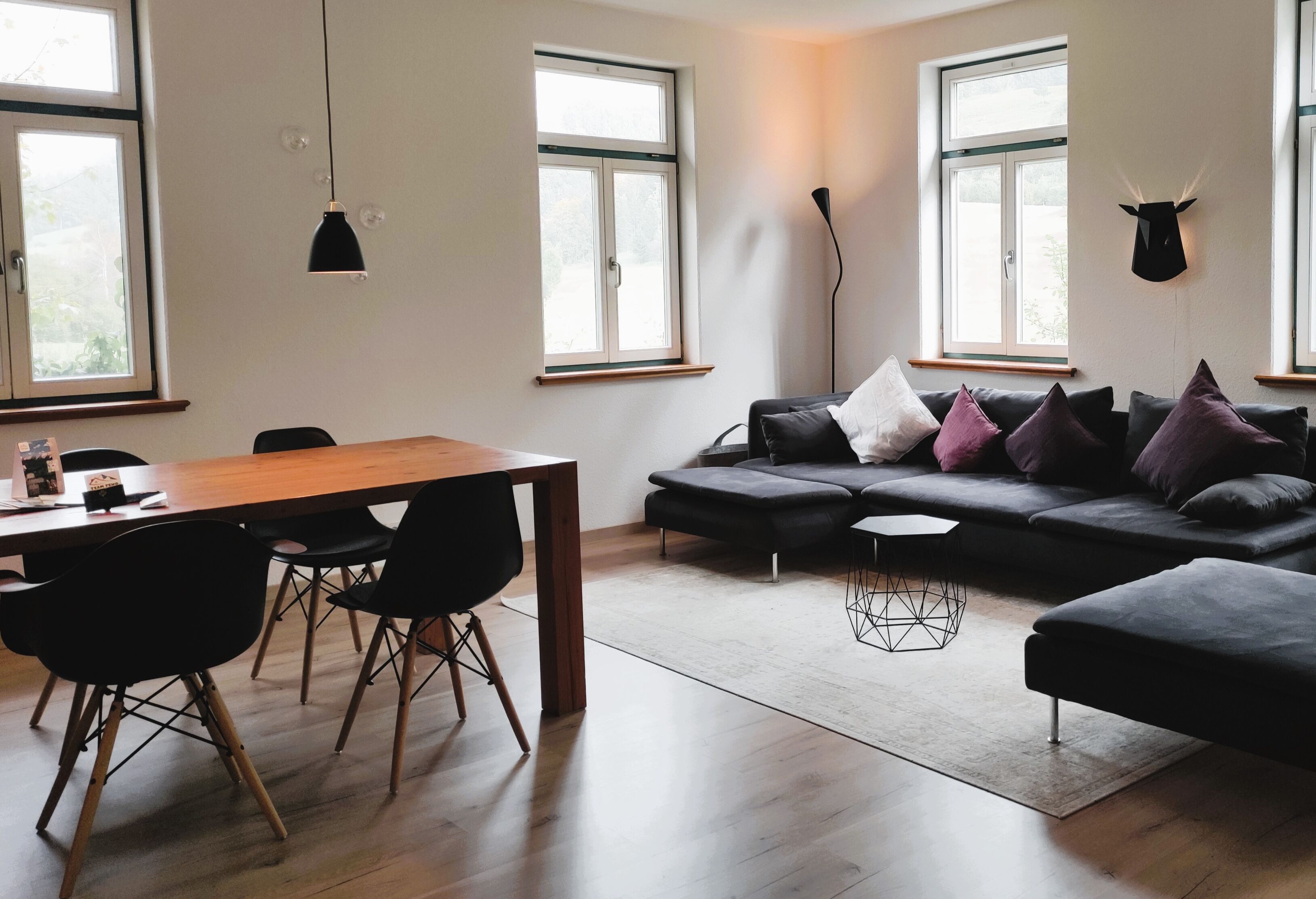 A modern living room with a wooden dining table, black chairs, a black sofa with cushions, and a large window with a bull's head decoration.