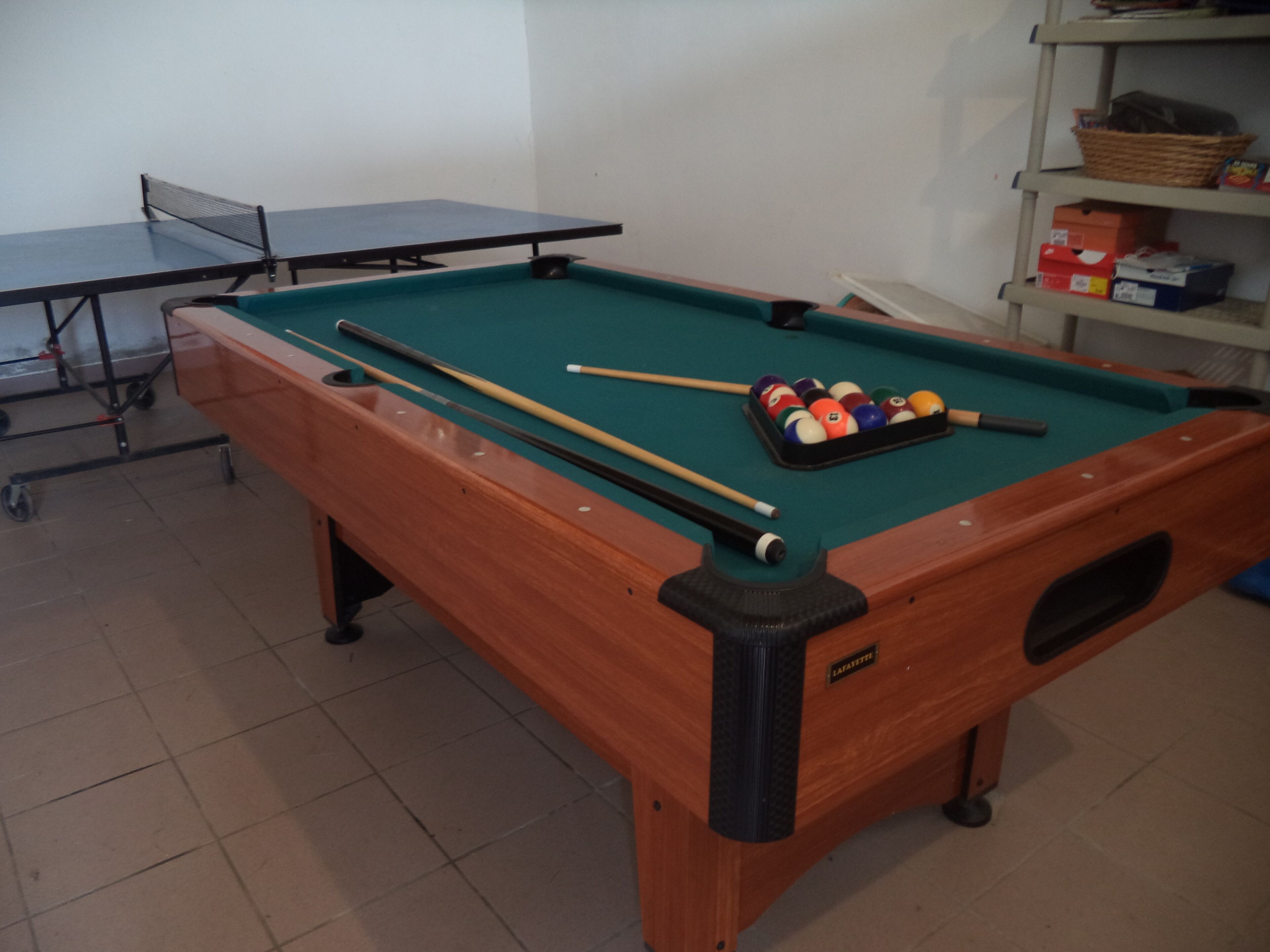 Une table de billard avec des billes et des queues, une table de ping-pong et une étagère avec des boîtes.