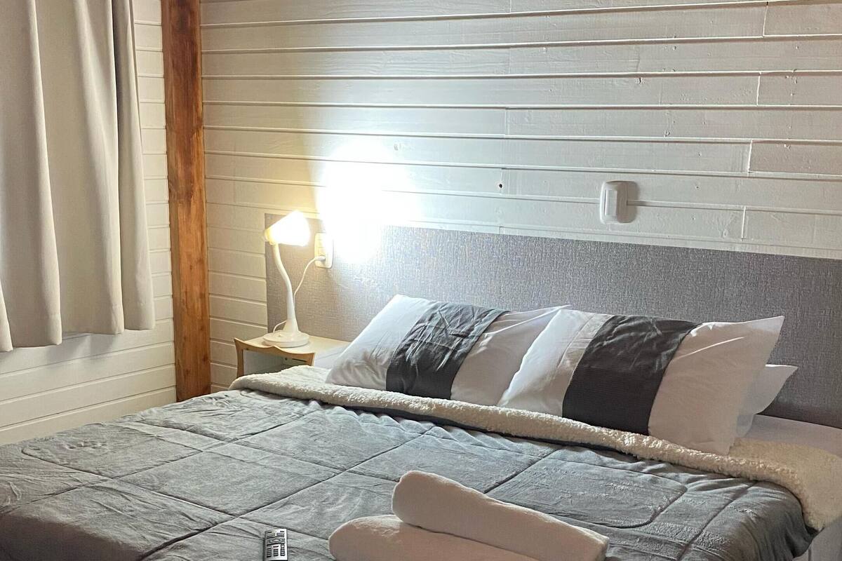 A neatly made bed with a grey quilt, two rolled towels, and a remote control on the bed.