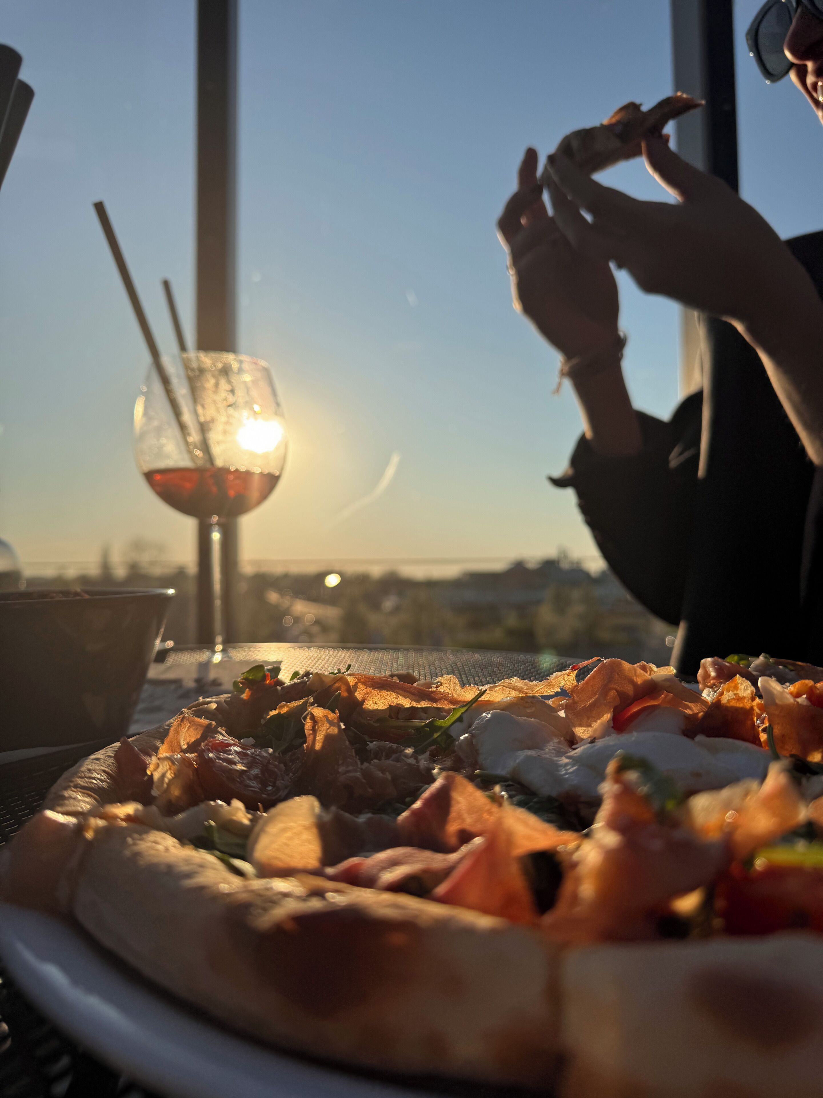 Une pizza sur une table, accompagnée d’un verre de vin rouge et d’une paille, le tout mis en valeur par une vue sur les gratte-ciel de la ville.