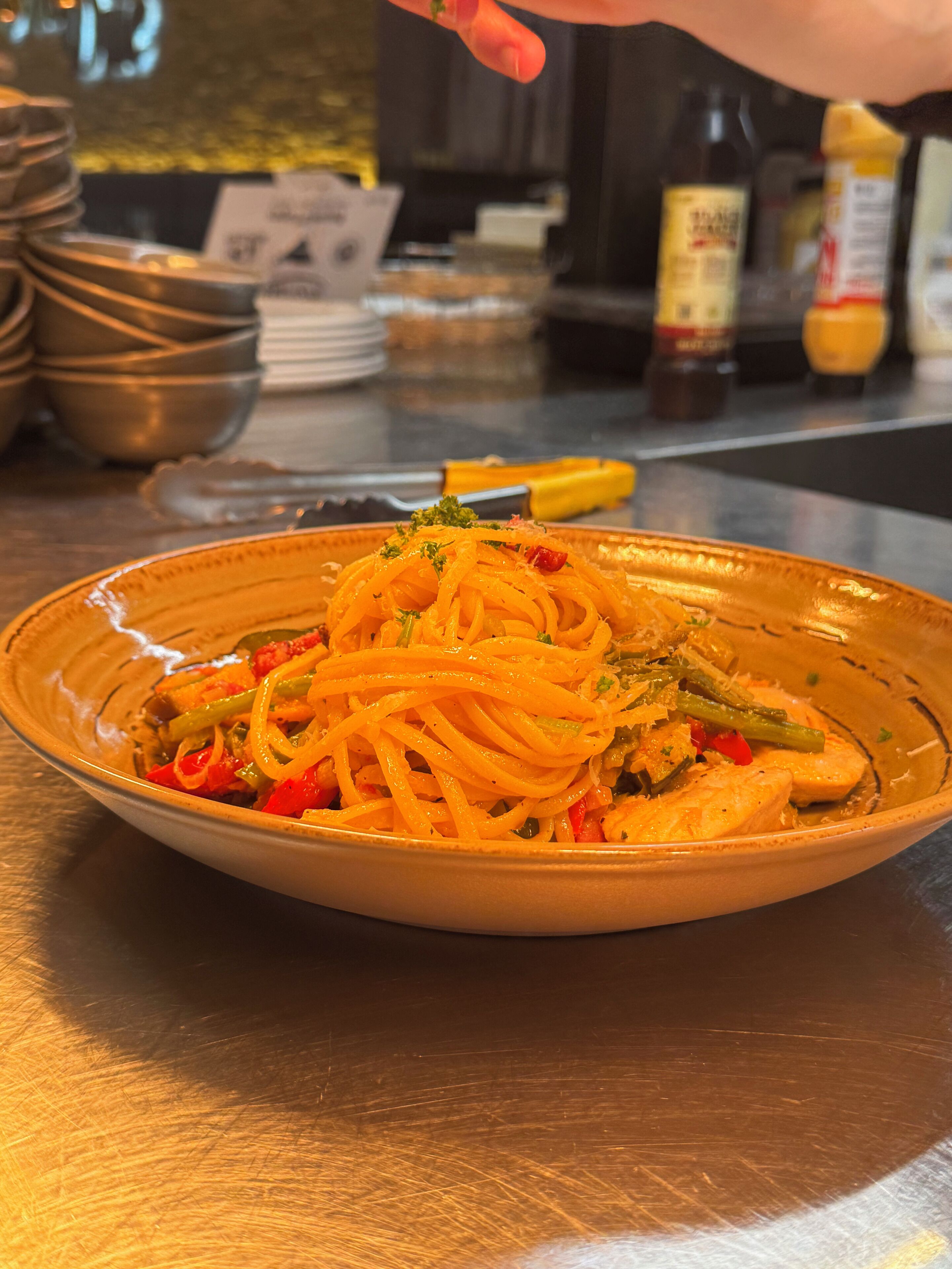 Un plato de espagueti con verduras y hierbas, sobre una mesa con utensilios de cocina y condimentos al fondo.