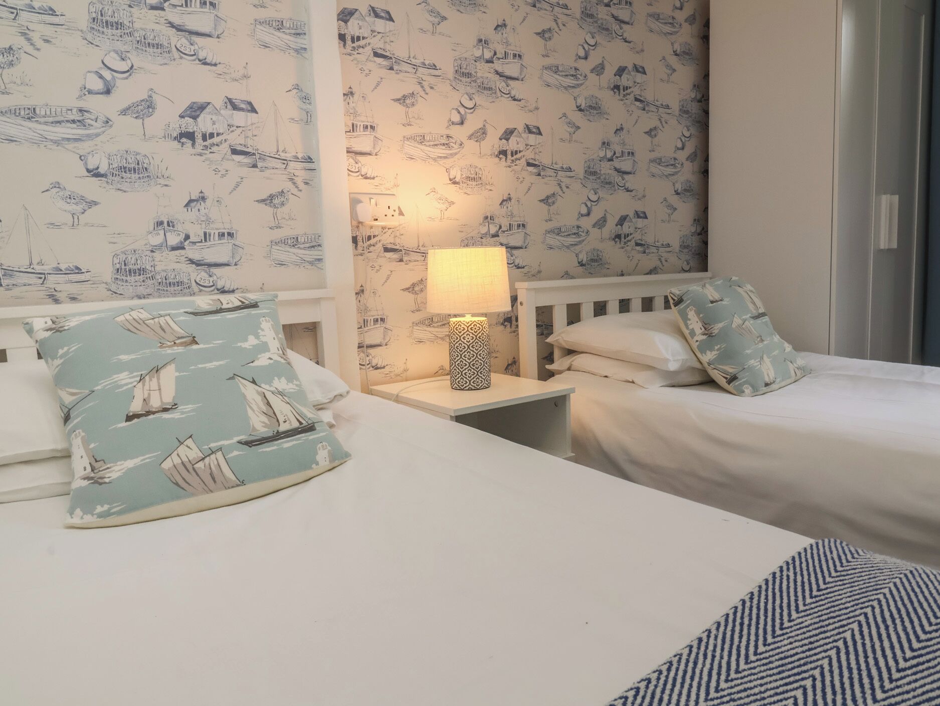 Habitación de hotel con dos camas, una mesita de noche con lámpara y papel tapiz con motivos náuticos.