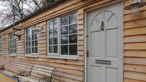 Un cottage in legno con una porta bianca con la scritta "Gatekeepers Cottage", una panchina e un cesto di fiori.