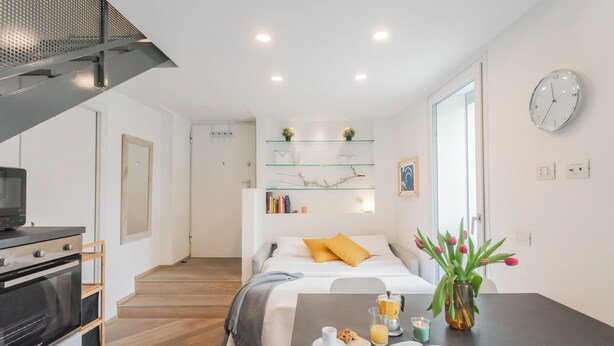 Un espacio habitable moderno y compacto con kitchenette, una cama con almohadas amarillas, una zona de comedor con mesa y una estantería.
