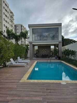A modern building with a pool, lounge chairs, and a patio area with plants.