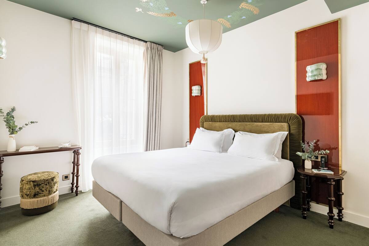 A neatly arranged hotel room with a large bed, a bedside table, a small stool, and a wall-mounted shelf.