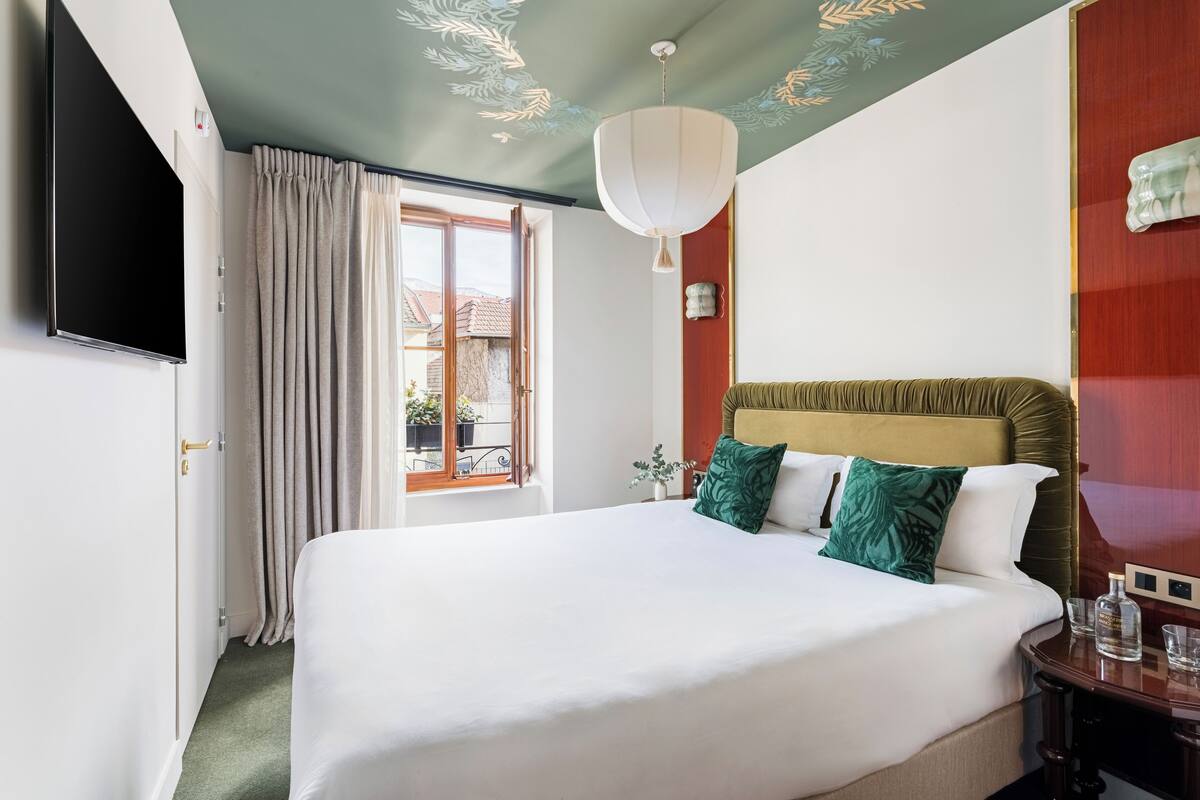 A hotel room with a large bed, a flat-screen TV, a window with curtains, and a chandelier.