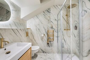 A bathroom with a marble wall, a glass shower enclosure, a gold showerhead, and a white sink. - La Cour du 6 (Annecy)