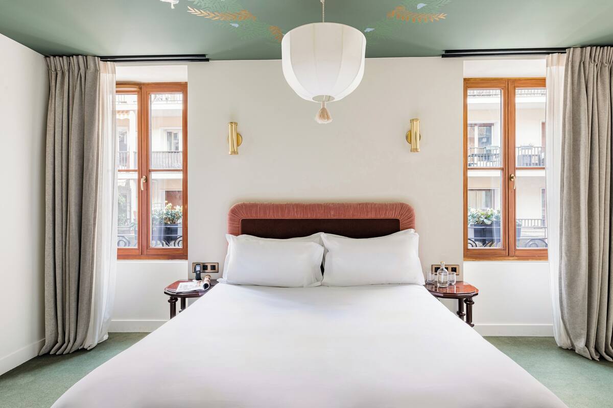 A hotel room with a large bed, a chandelier, two bedside tables, and windows with curtains.