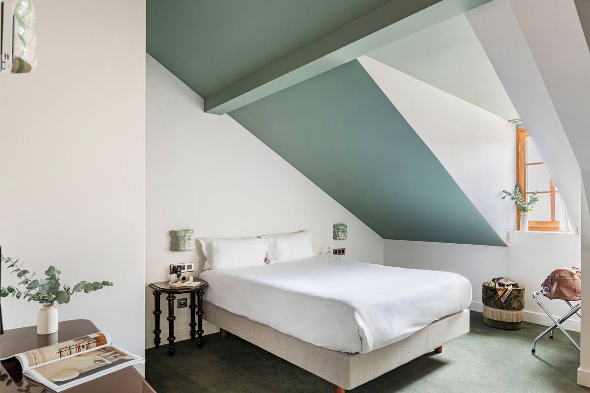 A bedroom with a sloped ceiling, a bed, a bedside table, a lamp, a plant, and a suitcase.