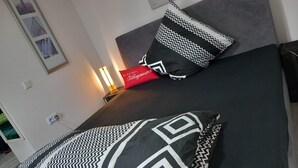 Ein Bett mit zwei geometrische Muster aufweisenden Kissen, ein rotes Kissen mit Text und eine an der Wand befestigte Lampe.
